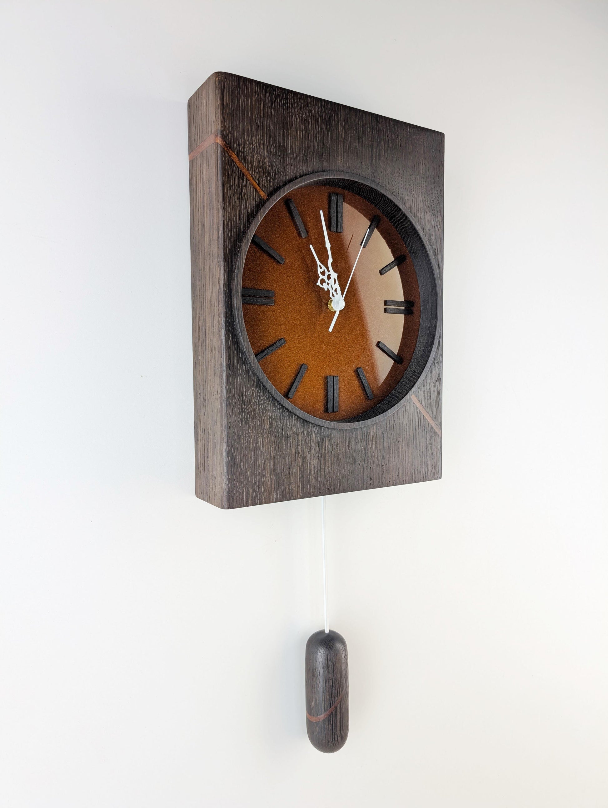 Schrägansicht der modernen Wanduhr Kupferzeit – Unikat aus geräucherter Eiche mit diagonalem Kupferharz-Inlay und Pendel, gefertigt von Steigerwood